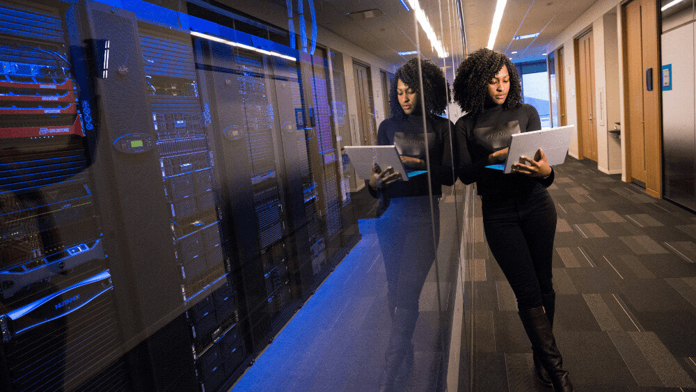 Photo of a woman working on a cybersecurity project. She is leaning on a window with a laptop in her hand.